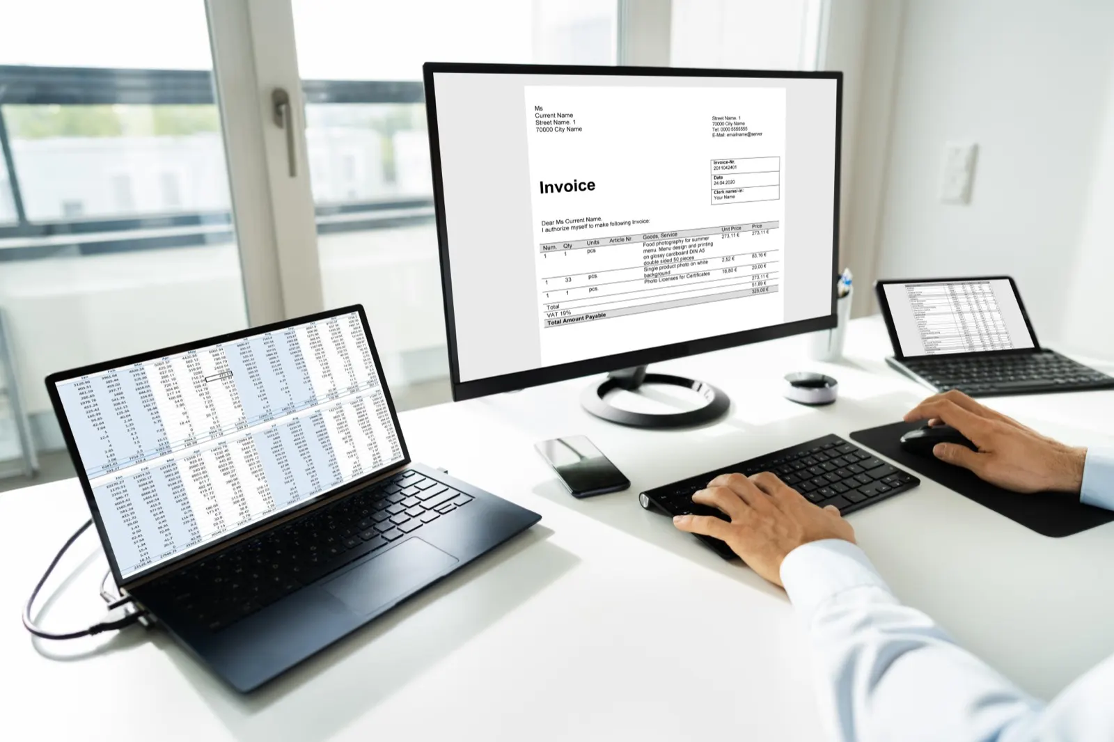 Ruky na klávesnici pri monitore s otvorenou faktúrou, vedľa notebook s tabuľkou