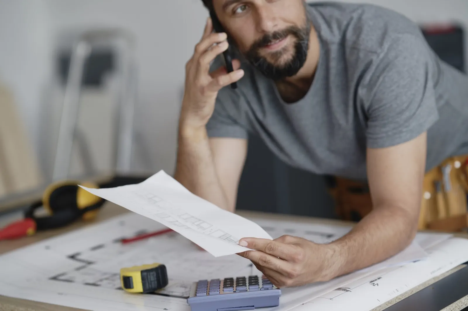 Remeselník opretý o pracovný stôl s plánmi telefonuje a drží v ruke papier